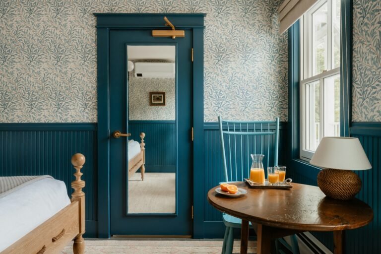 A bedroom with petrol-blue panelling, a blue door and window frames, small-print leafy wallpaper above, and a round wooden table set with orange juice beside a turned wooden bed