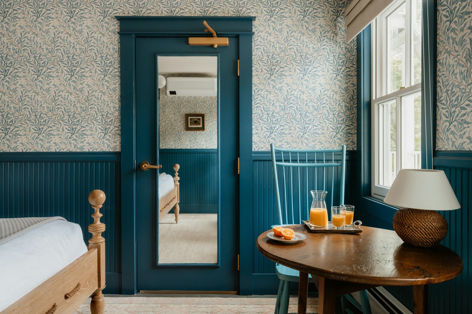 A bedroom with petrol-blue panelling, a blue door and window frames, small-print leafy wallpaper above, and a round wooden table set with orange juice beside a turned wooden bed