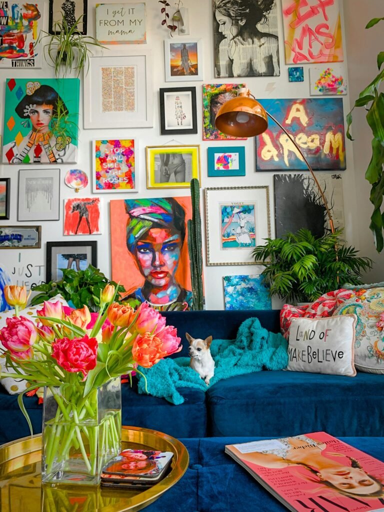 A wall packed with colourful framed art and neon paintings above a navy velvet sofa, with a chihuahua on a teal throw, patterned cushions and a vase of pink and orange tulips on a gold side table