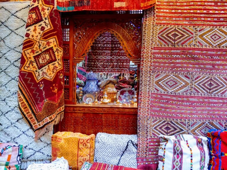 Stacks and hangings of patterned Moroccan rugs in red, orange, cream and black around a carved wooden alcove, with embroidered cushions piled on the floor
