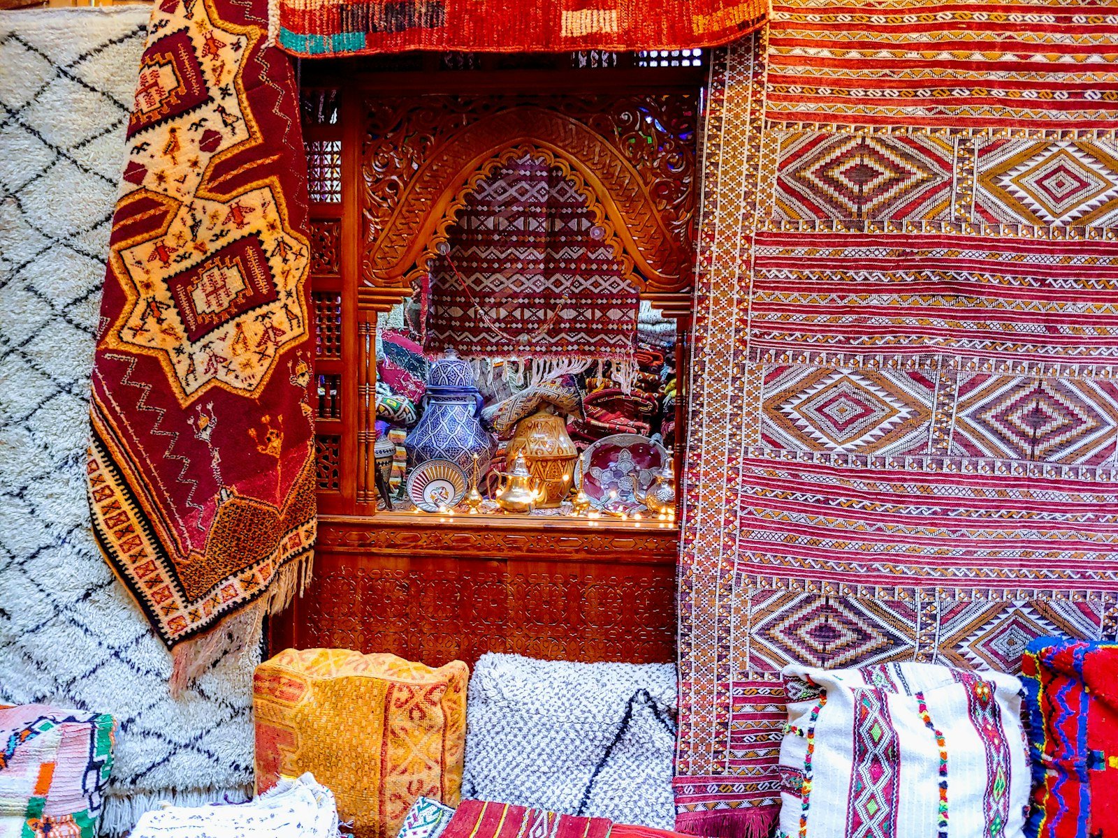 Stacks and hangings of patterned Moroccan rugs in red, orange, cream and black around a carved wooden alcove, with embroidered cushions piled on the floor