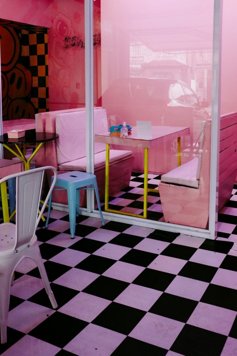 A hot-pink room with a black and white checkerboard floor, white bench seating, a pale blue stool and a yellow-legged table behind a glass partition