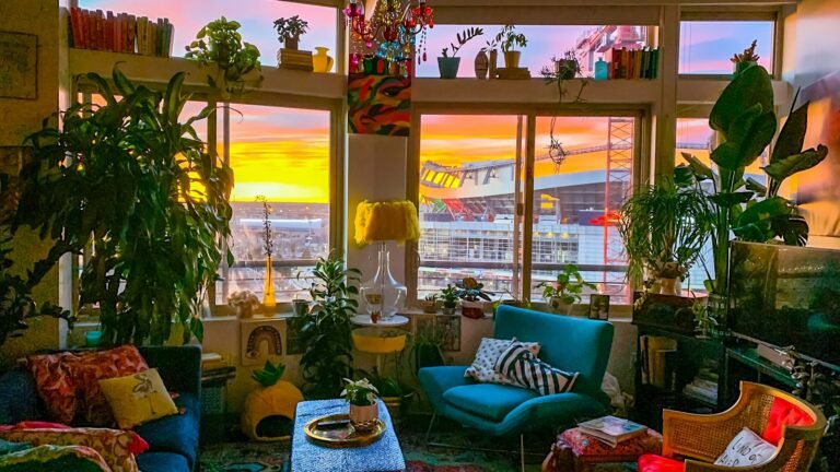A living room crowded with tall houseplants, a teal armchair, patterned cushions and a coffee table, in front of big windows glowing orange and pink at sunrise
