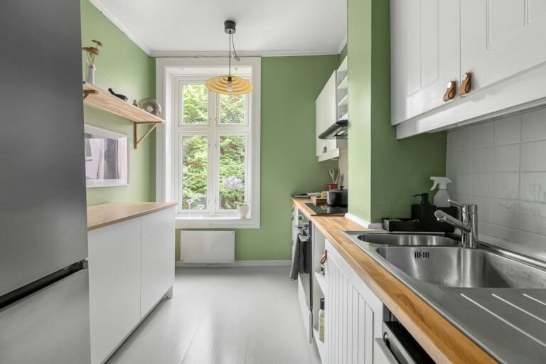 A narrow galley kitchen with sage green walls, white cabinets, a wooden worktop and a rattan pendant light in front of a tall window