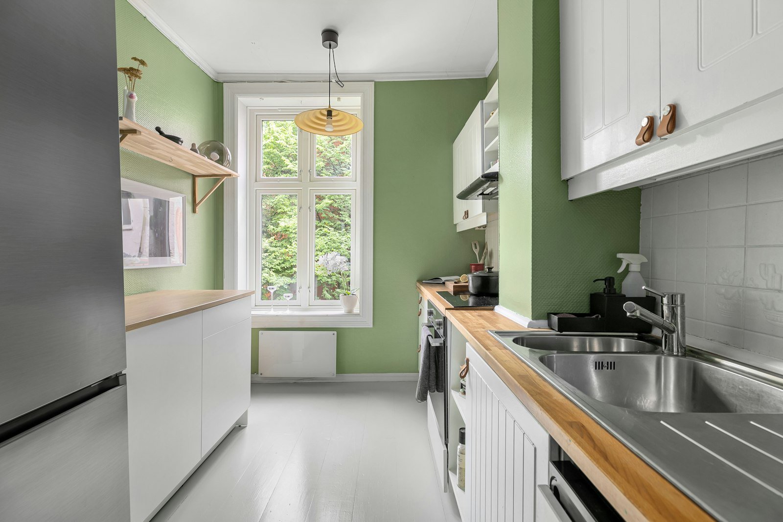 A narrow galley kitchen with sage green walls, white cabinets, a wooden worktop and a rattan pendant light in front of a tall window